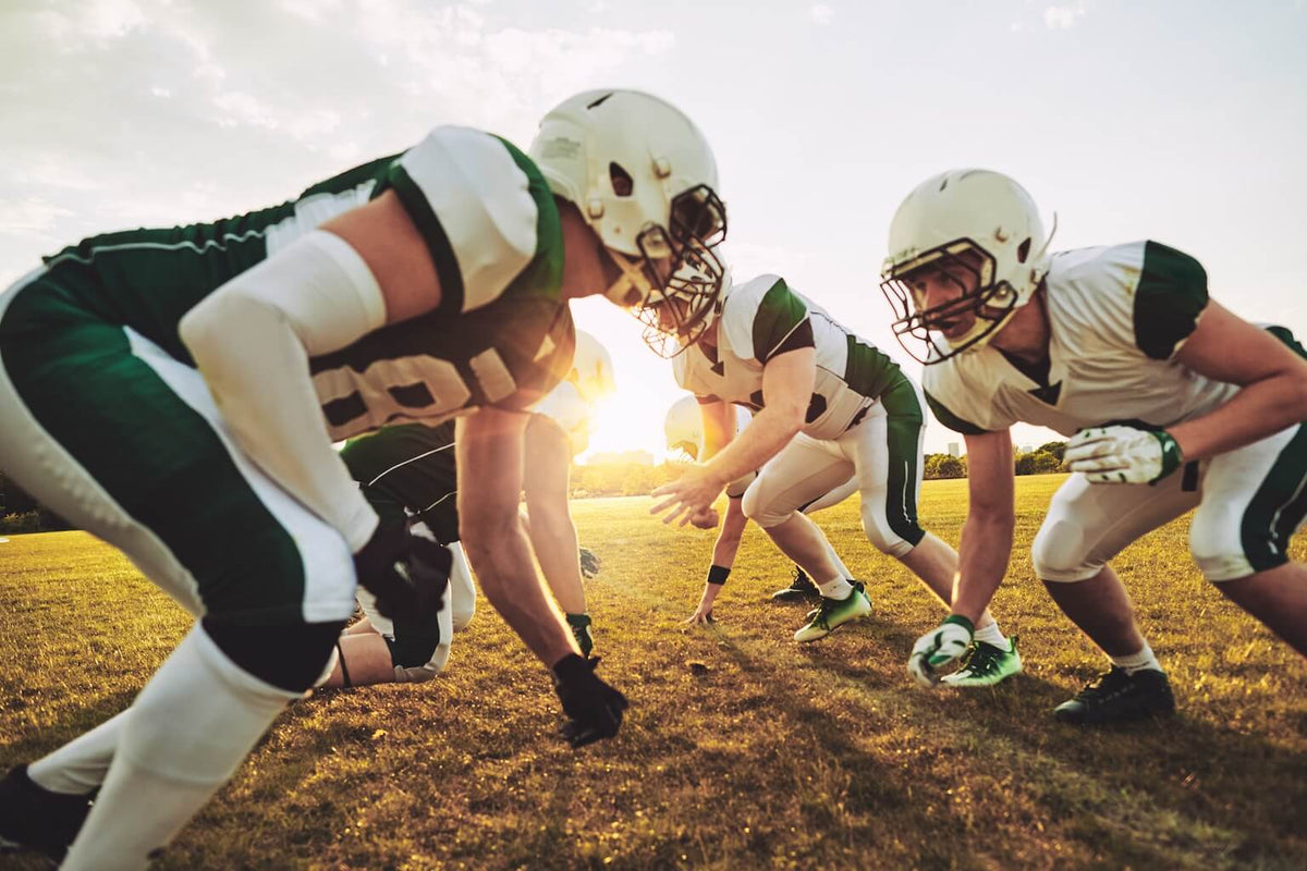 Two teams of football players in green and white uniforms face each other on a grassy field during a game, with the sun setting in the background.