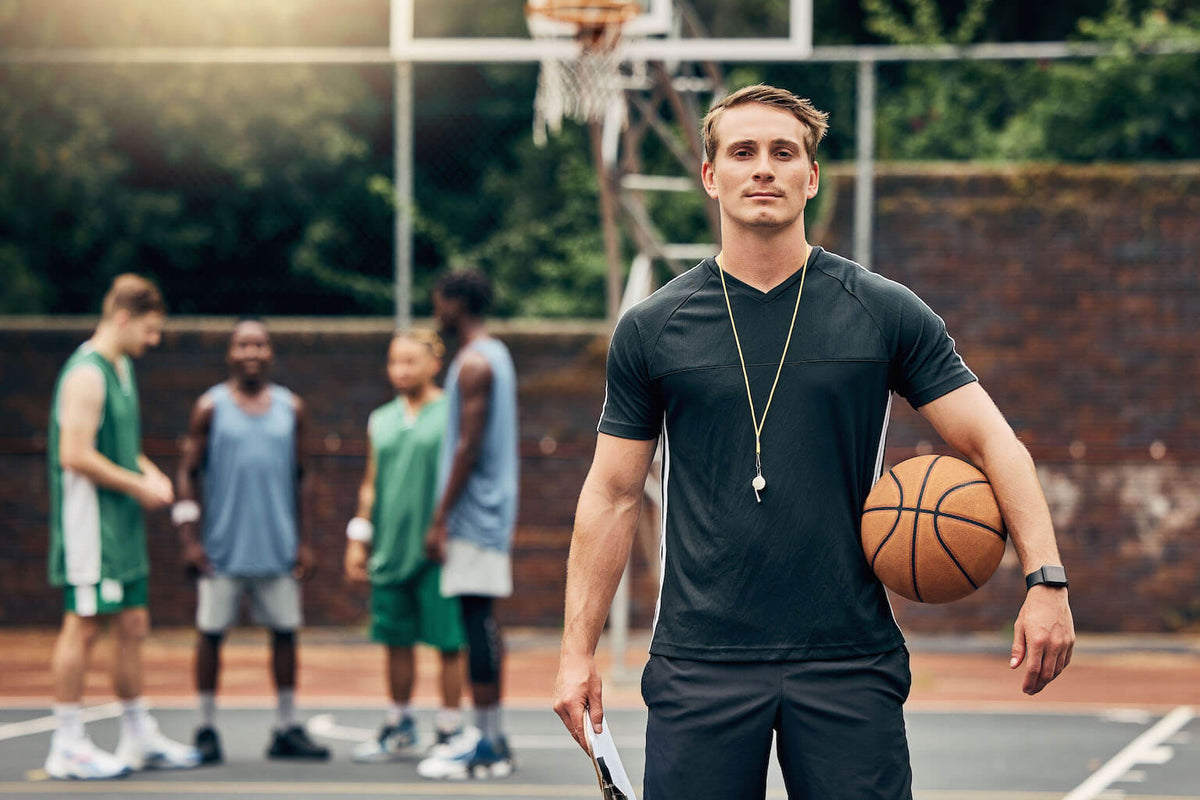 holding a basketball stands confidently on an outdoor court, with four other players in conversation in the background.