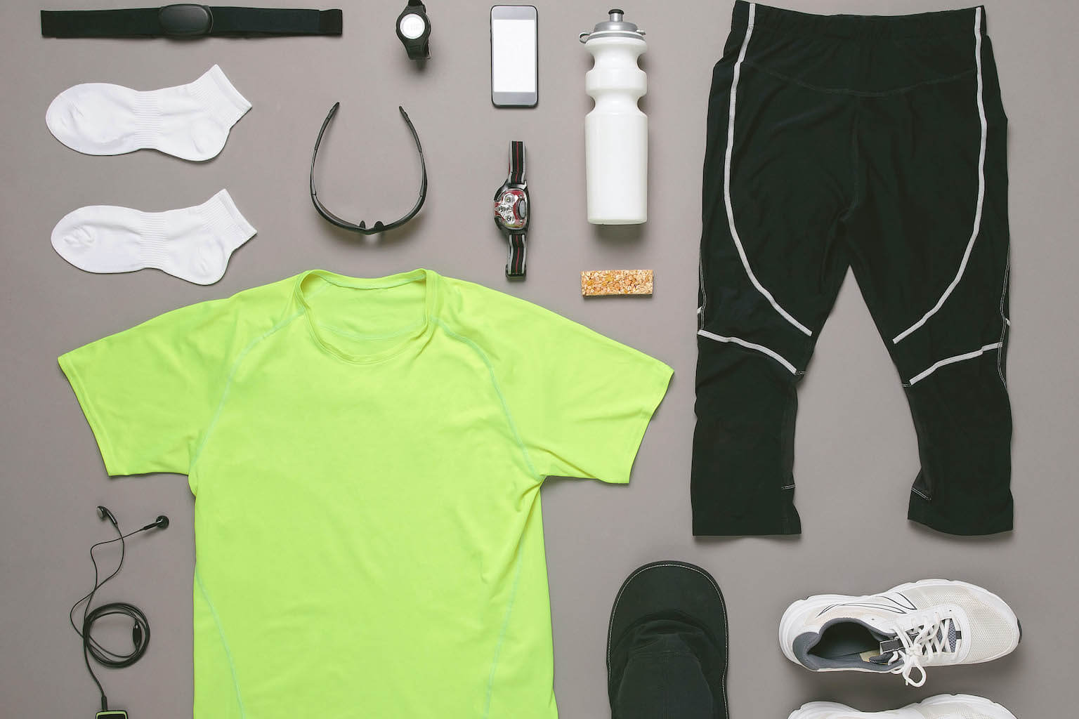Flat lay of jogging attire and accessories including a neon green t-shirt, black track pants, sneakers, headphones, a smartwatch, a water bottle, and a nutrition bar on a gray background.