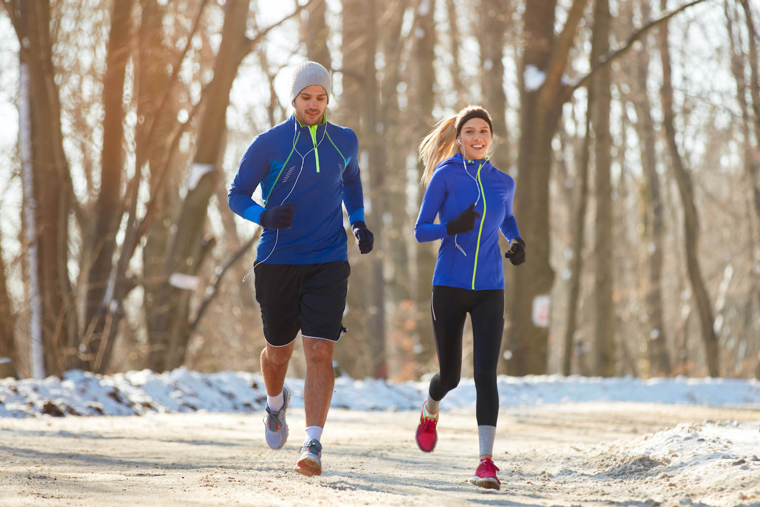 A man and a woman are jogging on a snow-lined path in a wooded area, wearing blue athletic gear and gloves.