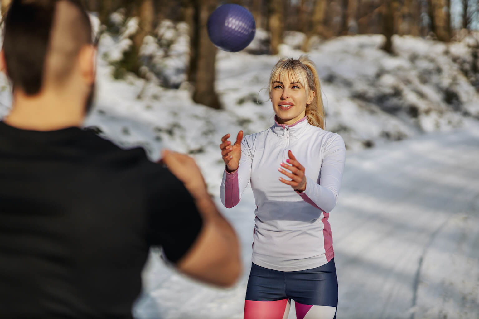 Woman catching purple ball in snowy landscape during winter training, with blurred man in foreground.