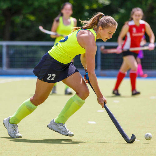 A female hockey player in neon green jersey taking the ball forward with her hockey stick