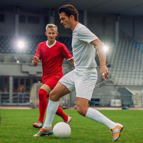 A soccer player in white jersey is kicking the ball while another player in an red jersey is trying to dodge