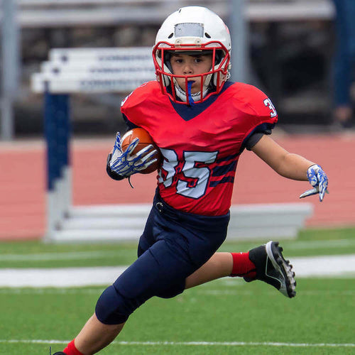 A boy in an red football jersey and white helmet, running while holding the ball in one hand