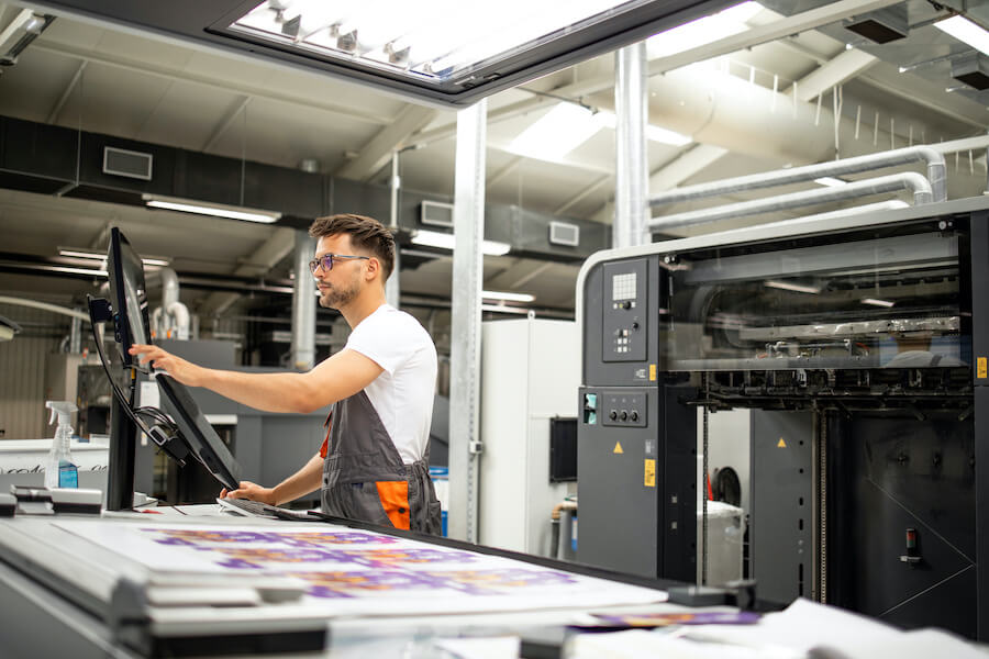 An employee looking into the monitor, standing beside the prints on the table with a factory equipment just behind him