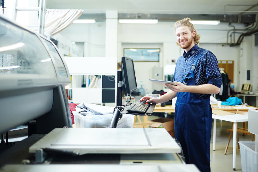 A smiling engineer in blue uniform, holding a tablet on hand and operating a huge print equipment